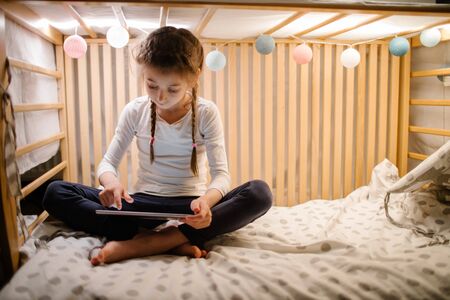 Child watching tablet pc at home. Girl watching a tablet and sitting in bed with garlands and having funの写真素材