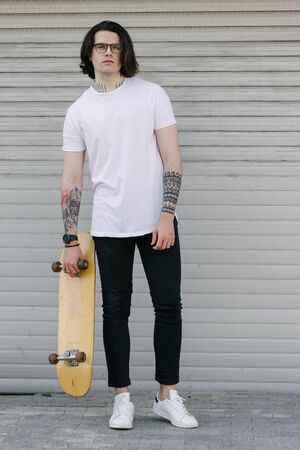 Hipster handsome male model wearing white blank t-shirt with space for your logo or design in casual urban style. He is holding a skateboardの写真素材