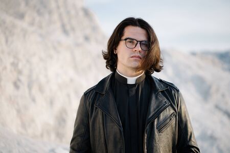Handsome young priest portrait outside at sunset. He is wearig eye glasses and roman collar shirt and a leather jacket.の写真素材