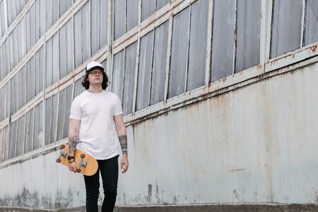 Handsome male model with beard wearing white blank t-shirt and a baseball cap with space for your logo or design in casual urban style. He is holding a skateboardの写真素材