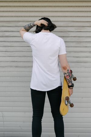 Hipster handsome male model wearing white blank t-shirt with space for your logo or design in casual urban style. He is holding a skateboardの写真素材