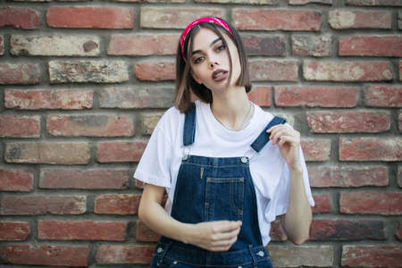 Slim sexy brunette girl wearing woman's denim overall over green tree wall texture in urban areaの写真素材