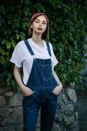 Slim sexy brunette girl wearing woman's denim overall over green tree wall texture in urban areaの写真素材