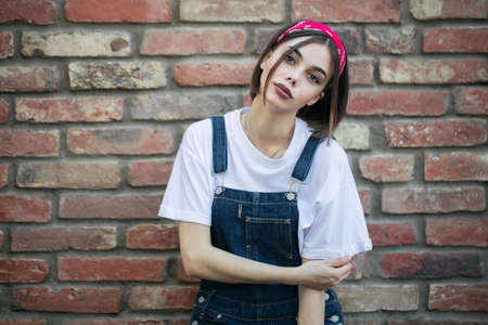 Slim sexy brunette girl wearing woman's denim overall over green tree wall texture in urban areaの写真素材