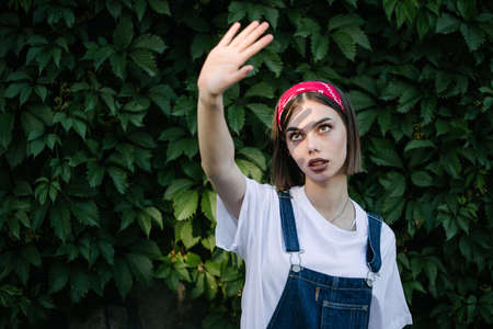 Slim sexy brunette girl wearing woman's denim overall over green tree wall texture in urban areaの写真素材