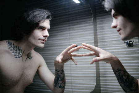 Young man in bathroom looking into a mirror at himself. He has a tattoo over his neckの写真素材