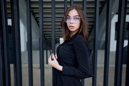 Business woman portrait in city streets at sunset. Sexy brunette wearing black dress, glasses and black leather bag outdoorの写真素材