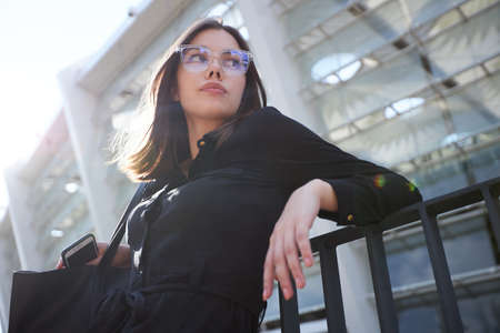 Business woman portrait in city streets at sunset. Sexy brunette wearing black dress, glasses and black leather bag outdoorの写真素材