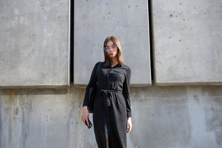 Business woman portrait in city streets at sunset. Sexy brunette wearing black dress, glasses and black leather bag outdoorの写真素材