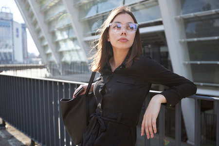 Business woman portrait in city streets at sunset. Sexy brunette wearing black dress, glasses and black leather bag outdoorの写真素材