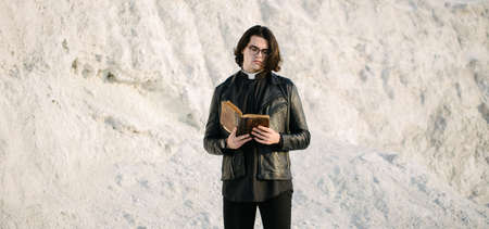 Young priest portrait outside at sunset. He is wearig eye glasses and roman collar shirt and a leather jacket.の写真素材