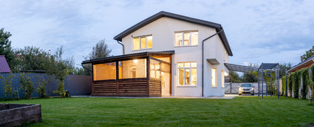 Exterior view of modern white house with courtyard, patio area, green grass lawn, garden and a car in the eveningの写真素材