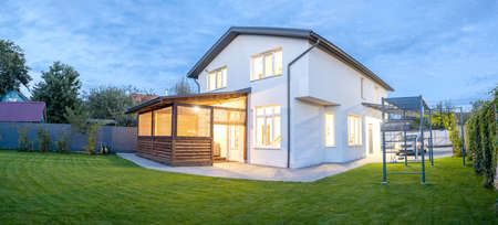 Exterior view of modern white house with courtyard, patio area, green grass lawn, garden and a car in the eveningの写真素材