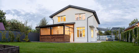 Exterior view of modern white house with courtyard, patio area, green grass lawn, garden and a car in the eveningの写真素材