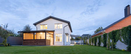 Exterior view of modern white house with courtyard, patio area, green grass lawn, garden and a car in the eveningの写真素材