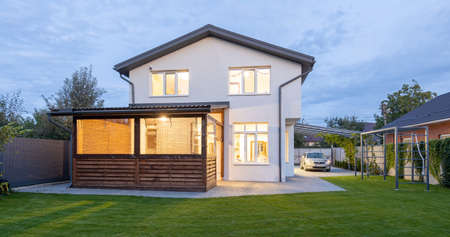 Exterior view of modern white house with courtyard, patio area, green grass lawn, garden and a car in the eveningの写真素材