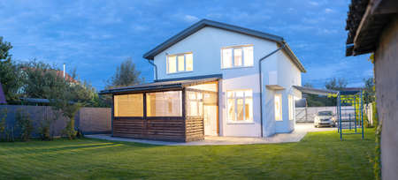 Exterior view of modern white house with courtyard, patio area, green grass lawn, garden and a car in the eveningの写真素材