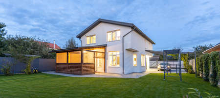 Exterior view of modern white house with courtyard, patio area, green grass lawn, garden and a car in the eveningの写真素材