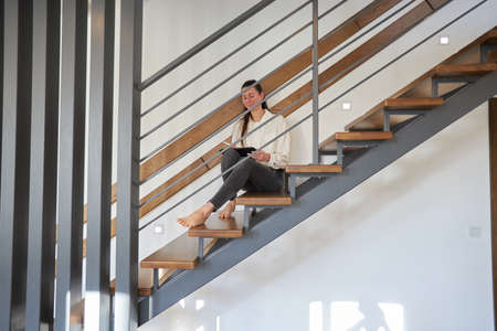 Woman working from home in her living room while sitting on staircase. Modern metal stairway in interior designの写真素材