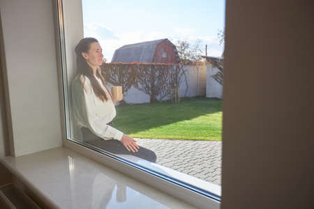 Woman relaxing in her backyard next to a window while drinking coffeeの写真素材