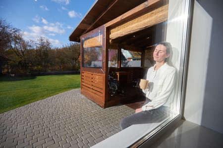 Woman relaxing in her backyard next to a window while drinking coffeeの写真素材