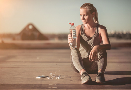 Attractive sporty girl is having rest and drinking water after the jogging on street. Smart phone with headphones are nearby.の写真素材