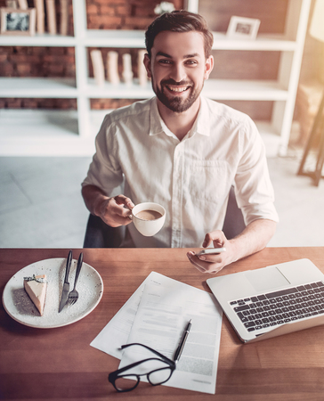 Handsome man is working in cafe, drinking cup of coffee, smiling and looking at camera.の写真素材