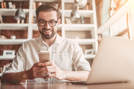 Handsome man in eyeglasses is working in cafe with smart phone in hands and smiling.の写真素材