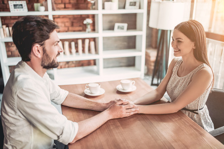 Couple in love is holding hands on wooden table and look on each other. Cups of coffee are on the background.の写真素材