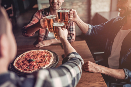 Cheers! Side view of three male friends in bar drinking beer and eating pizza.の写真素材