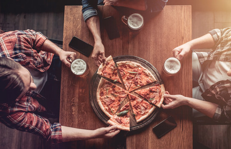 Top view of three male friends in bar drinking beer and eating pizza.の写真素材