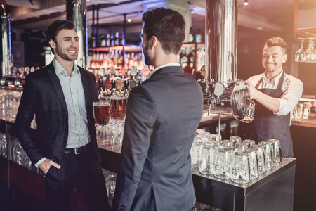 Handsome young businessmen are drinking beer, communicating and smiling. Bartender is on a background.の写真素材