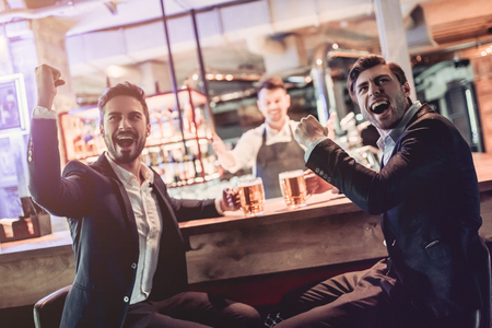 Handsome businessmen are sitting on a bar counter, drinking beer and watching football. Active fans.の写真素材