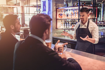Handsome businessmen in bar are drinking beer and communicating with cheerful bartender.の写真素材