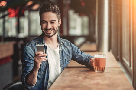 Portrait of handsome young man in bar drinking beer, looking at smartphone and smilingの写真素材