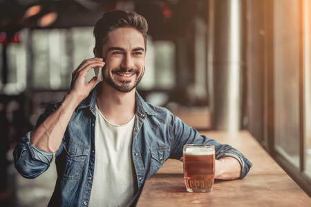 Portrait of handsome young man in bar drinking beer, talking on smartphone and smilingの写真素材