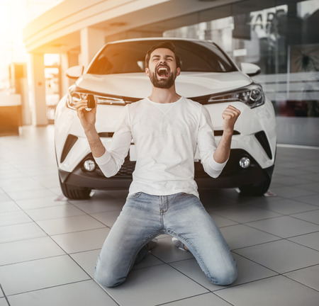 Yes, that's my new car! Customer in car dealership. Young man knelt down with keys in hand.の写真素材