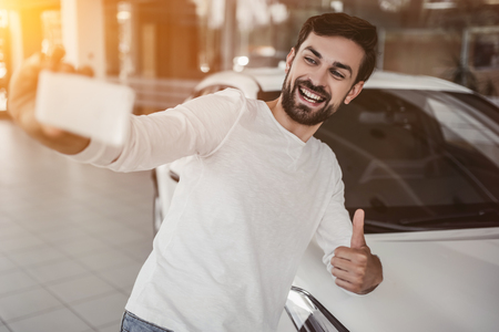 Young man is choosing a new vehicle in car dealership and making photo on a smartphone.