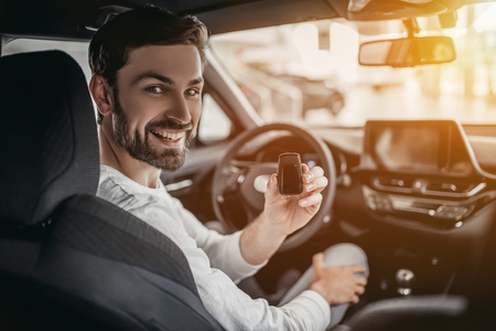 Young man is choosing a new vehicle in car dealership. Holding keys in hand, smiling and looking at the camera.の写真素材