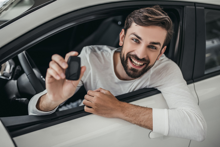 Young man is choosing a new vehicle in car dealership. Holding keys in hand, smiling and looking at the camera.の写真素材