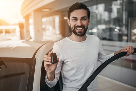 Young man is choosing a new vehicle in car dealership. Holding keys in hand, smiling and looking at the camera.の写真素材
