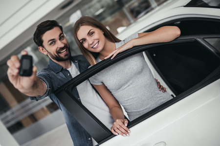 Happy beautiful couple is choosing a new car at dealership, smiling and looking at camera with keys in hand.の写真素材