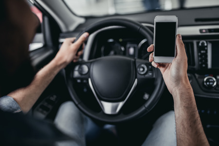 Cropped image of young man is sitting in new car at car dealership with smartphone in hand.の写真素材