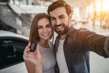 Happy beautiful couple is choosing a new car at dealership. Man is making photo while woman is taking keys in hand.の写真素材