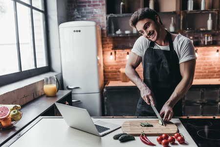 Attractive young man is cooking on kitchen with laptop on table while talking on smart phone.の写真素材