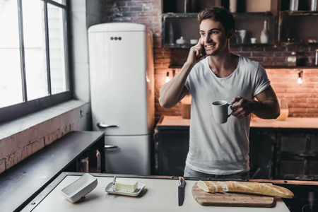 Handsome man on kitchen is smiling, drinking coffee and talking on a smart phone.の写真素材