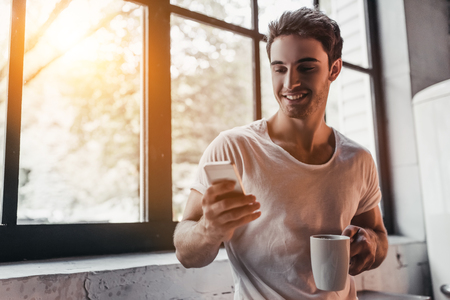 Handsome man on kitchen is smiling, drinking coffee in the morning and looking at smart phone.の写真素材