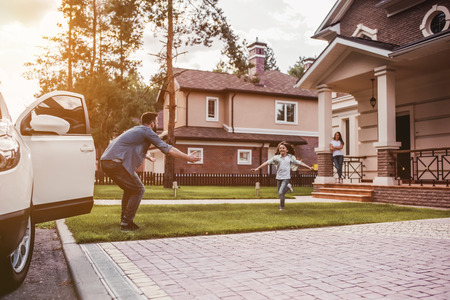 Happy family. Dad came home, daughter is running to meet him while wife is waiting on the house's porch.