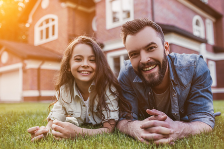 Dad and daughter are lying on the green grass and having fun on the backyard. Smiling and looking at camera.の写真素材