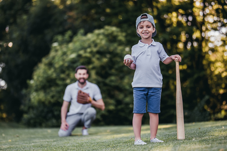 Handsome dad with his little cute sun are playing baseball on green grassy lawn